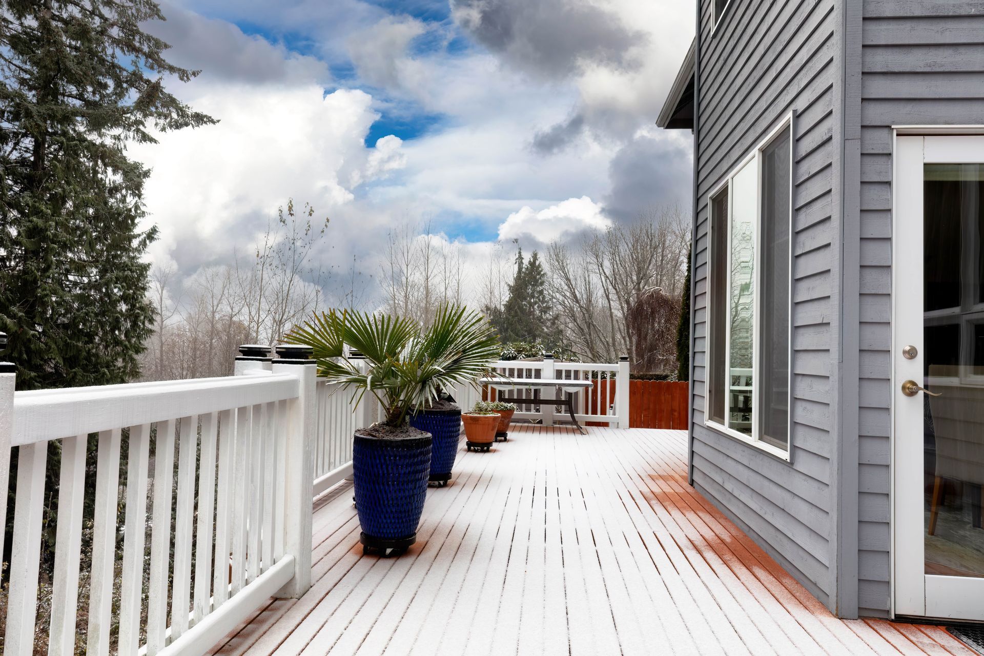 Wooden deck with potted plants, railing, and gray house against a cloudy sky.