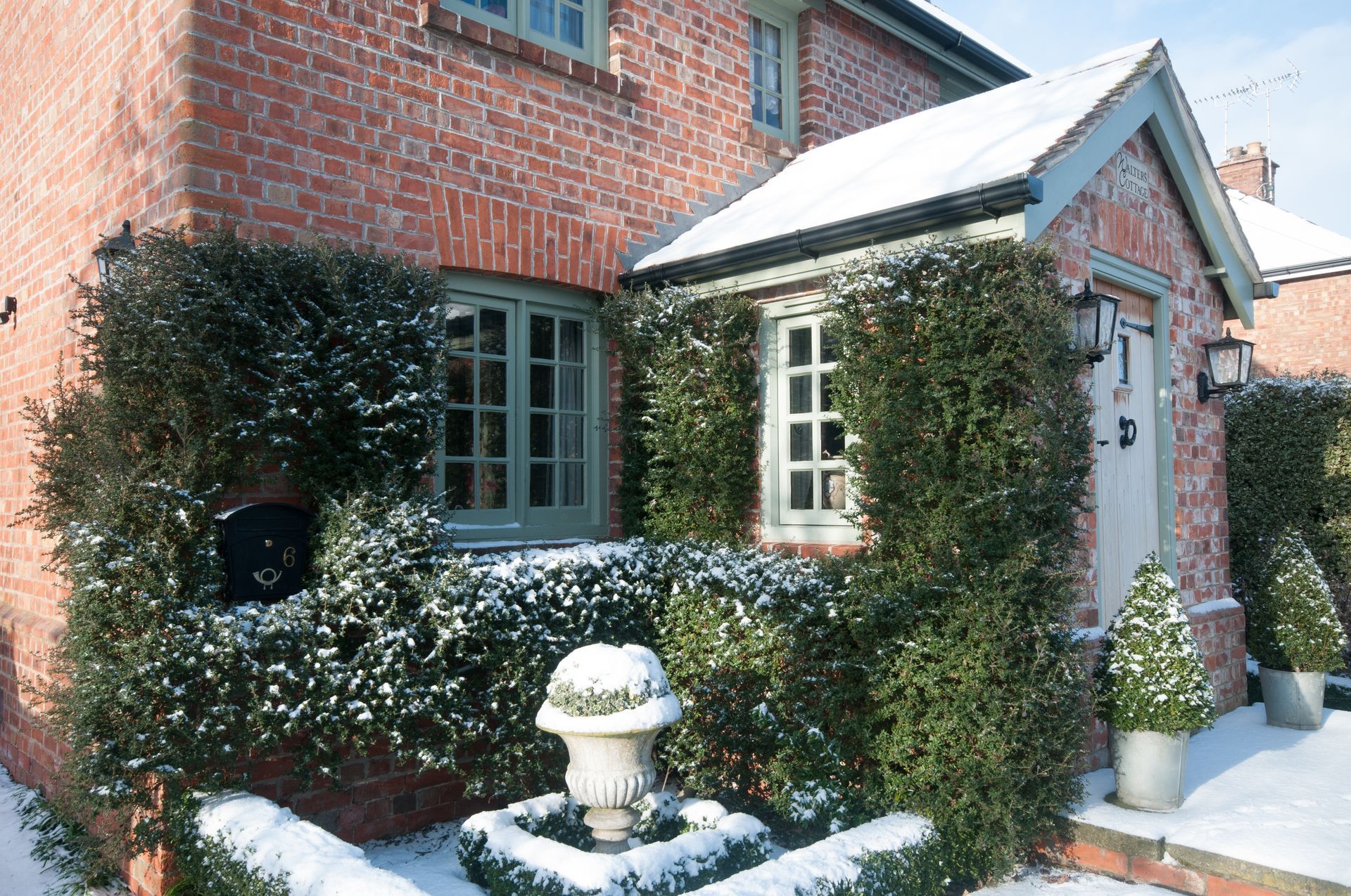 Brick cottage covered in snow, with green-framed windows and entrance, and a small fountain.