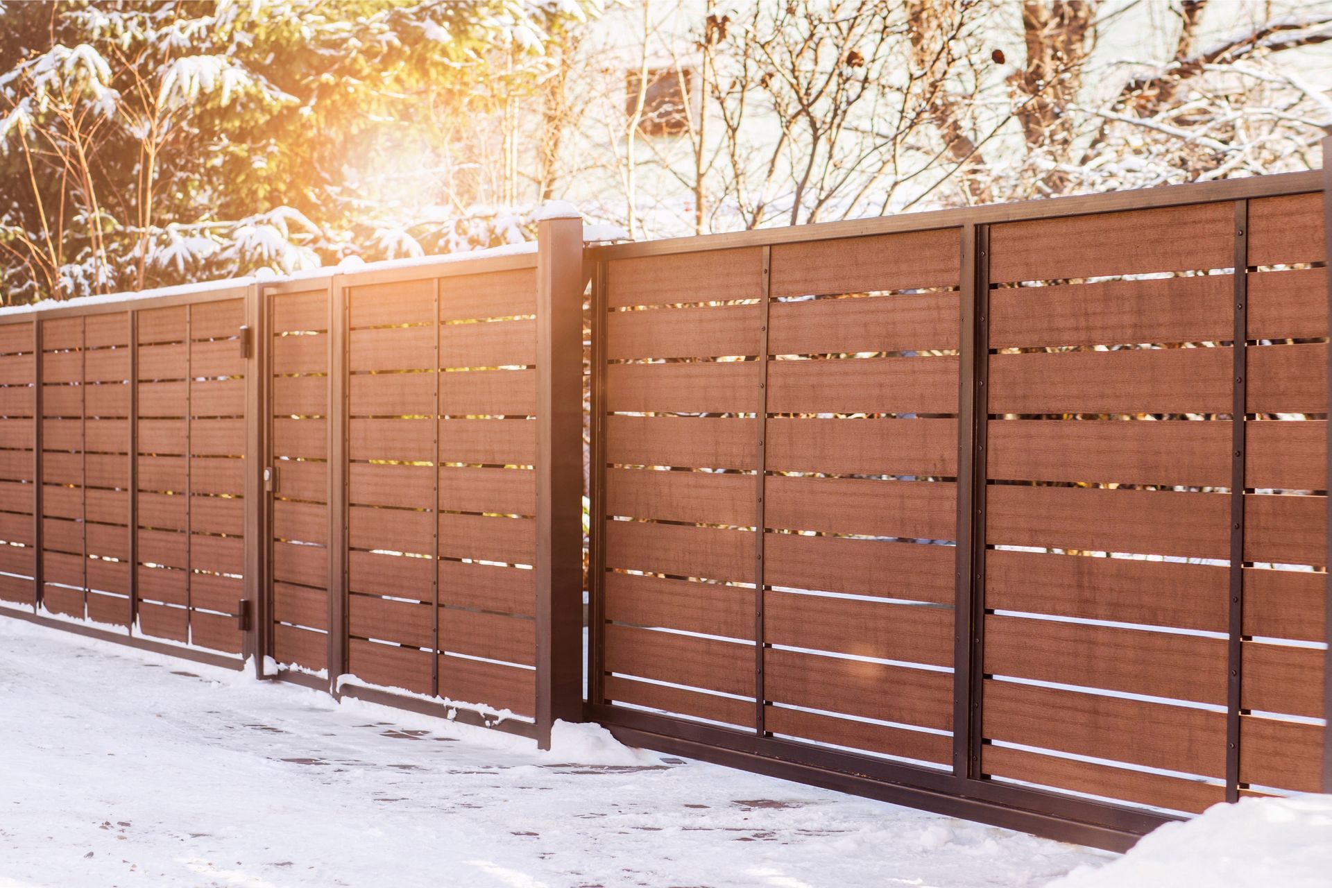 Brown slatted privacy fence with metal posts in a snowy outdoor setting, sunlit from the top left.