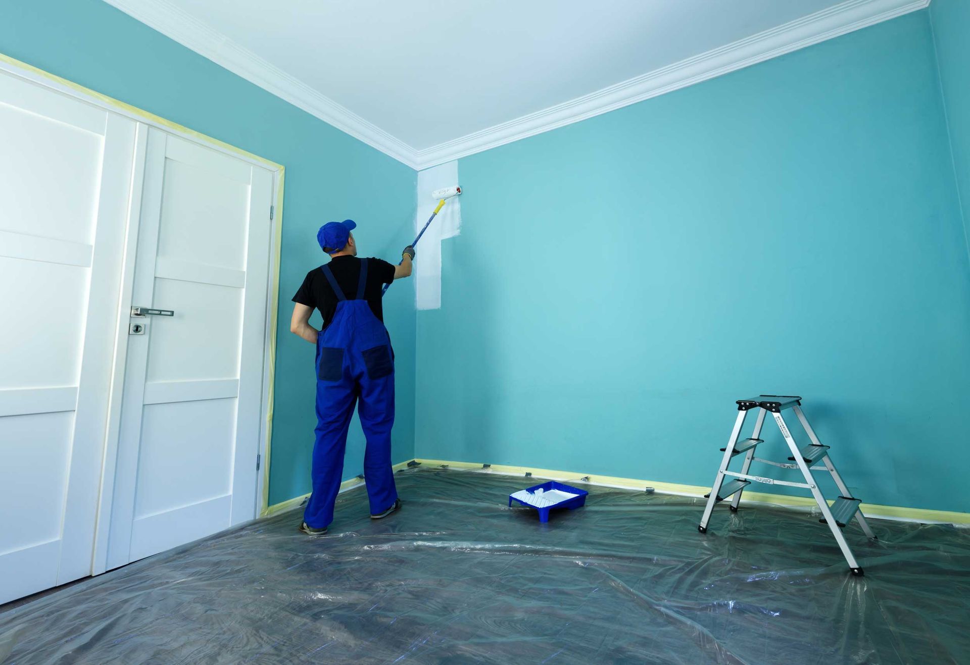 A man in working overalls is painting a wall in white color