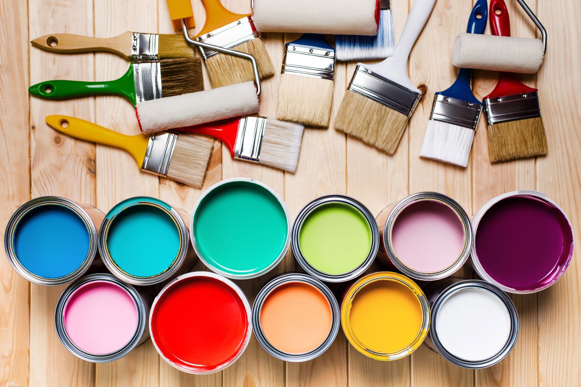 Paint cans filled with various colors with paint brushes and rollers on a wooden surface.