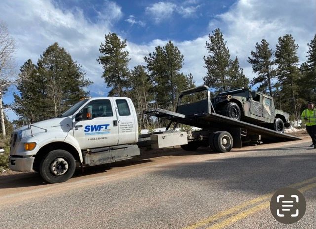 A tow truck with a military vehicle on the back is parked on the side of the road.