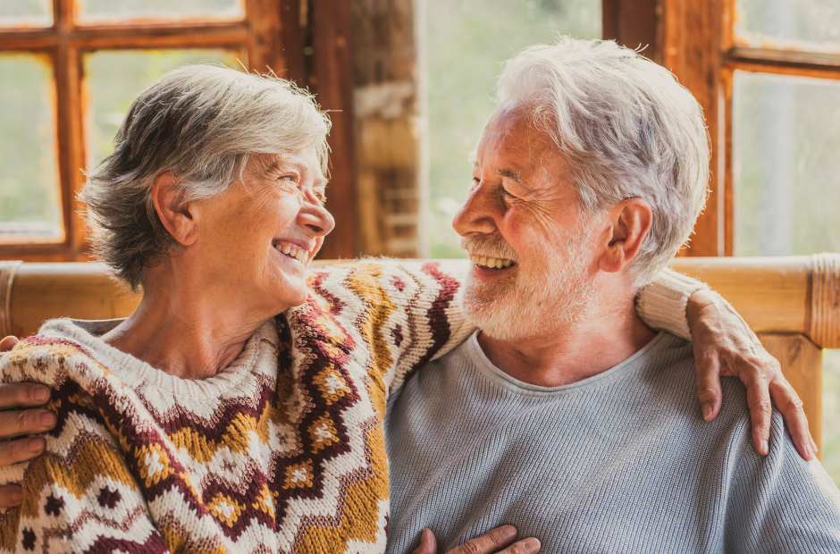 An elderly couple is sitting on a couch and smiling at each other.