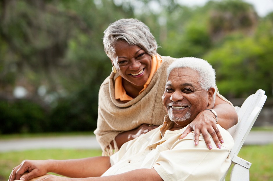 A woman is standing next to an older man sitting in a chair.