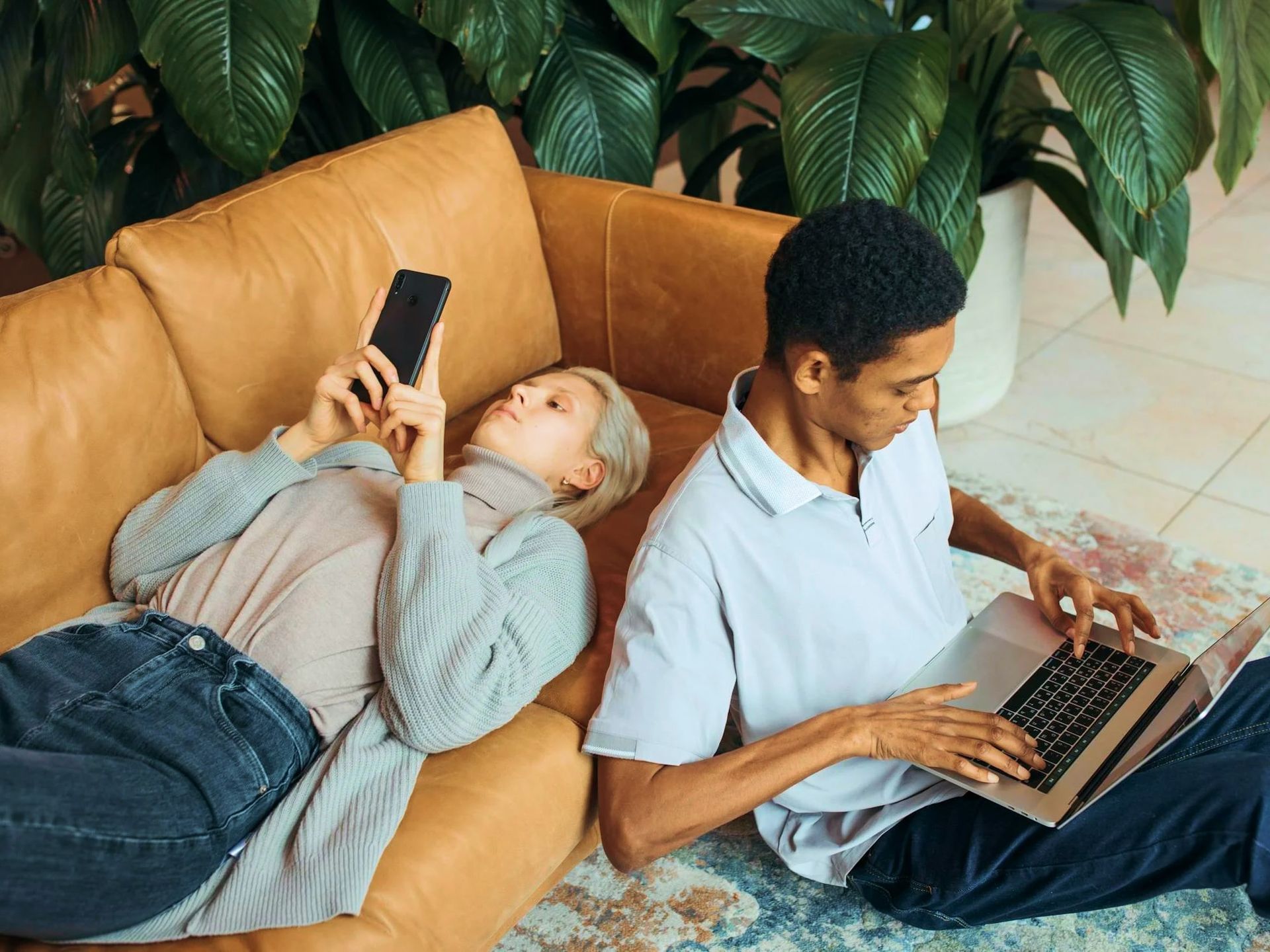 A person lying on a brown leather couch uses a phone, while another person sits on the floor nearby using a laptop.