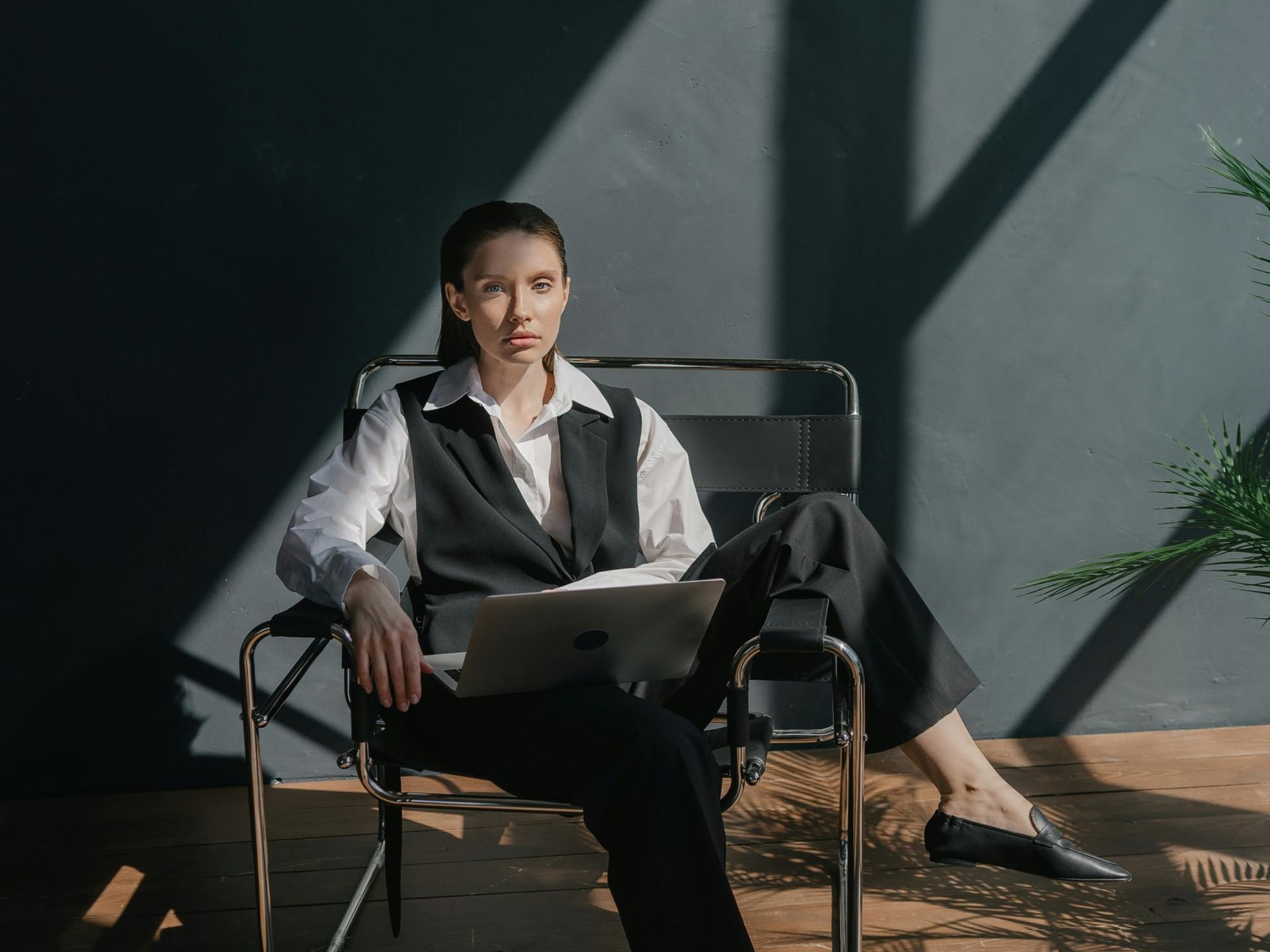 A person in professional attire sits in a modern chair holding a laptop, illuminated by sunlight against a dark wall.