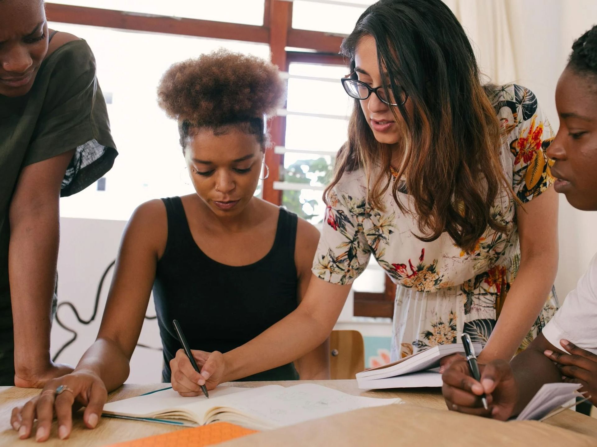 Four people collaborate around a table, writing in notebooks and pointing at documents in a brightly lit room.