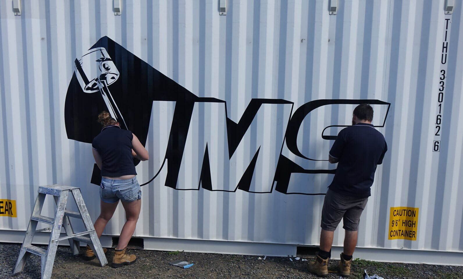 A Man And A Woman Are Painting The Word Umg On A Shipping Container — Sun City Signs In Paget, QLD