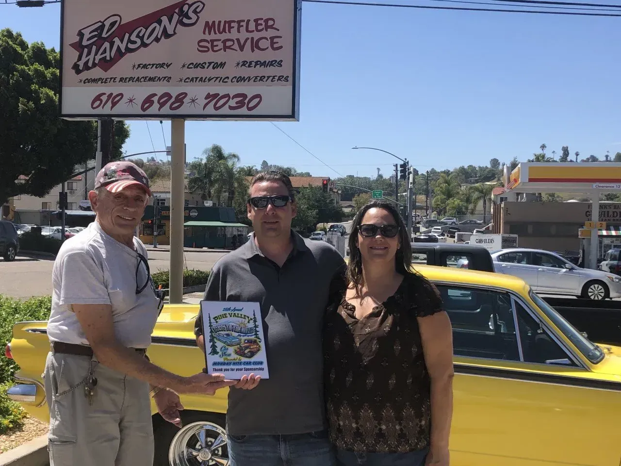 Three People Stand in Front of Ed Hanson's Muffler Service's Sign