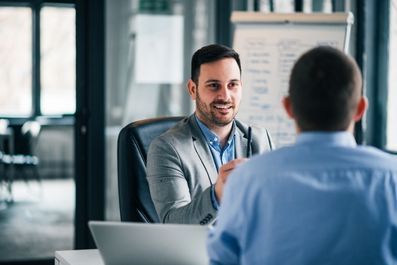 Man in blazer smiles, facing another person; office setting with a whiteboard and laptop.