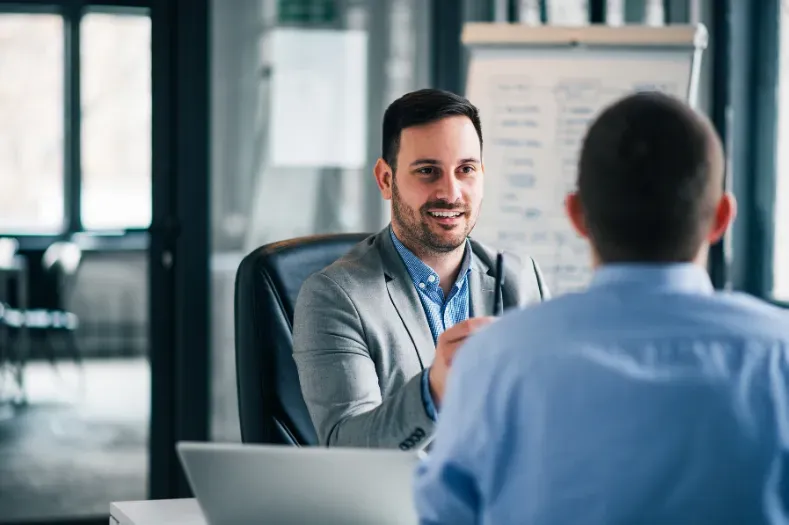Man in blazer smiles during interview. Another person in blue shirt faces him. Office setting.