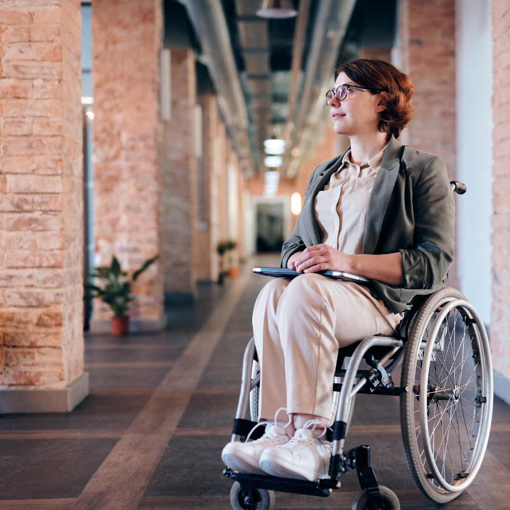 Woman in a wheelchair, looking towards the right, holding a tablet, in a brick-walled hallway.