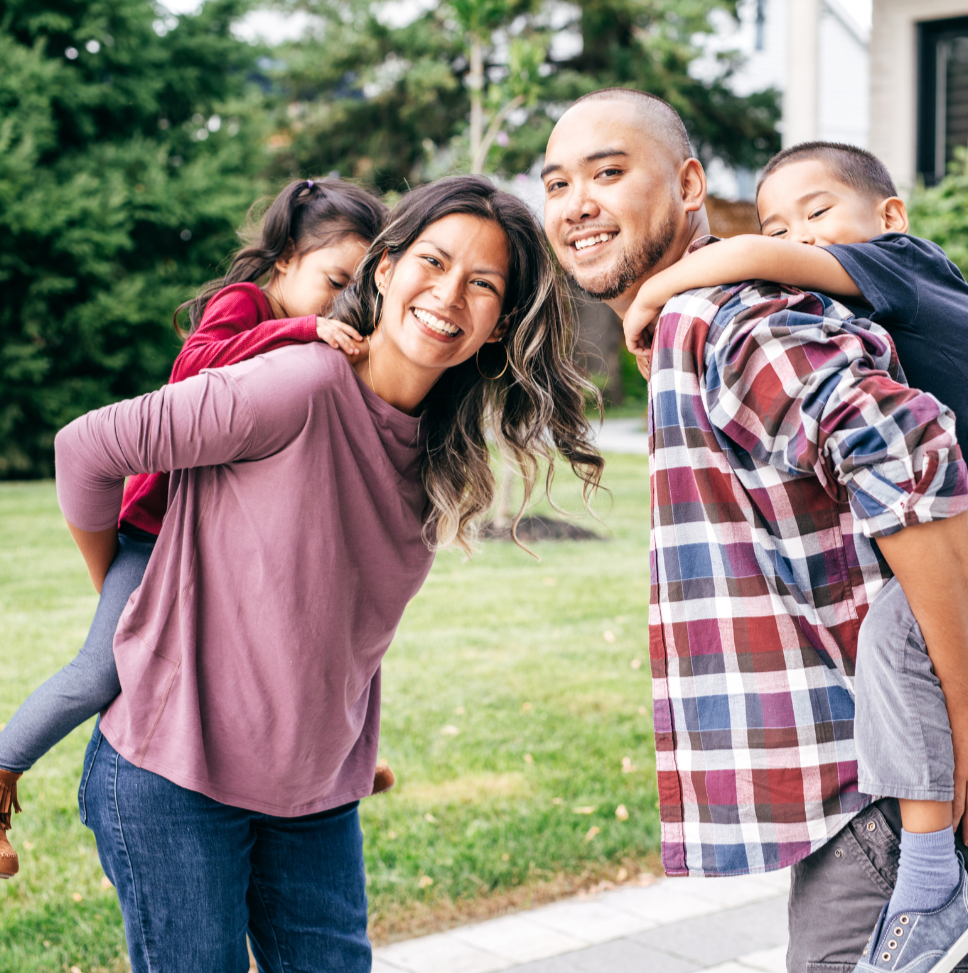 Family smiling while giving each other piggyback rides outside a house.