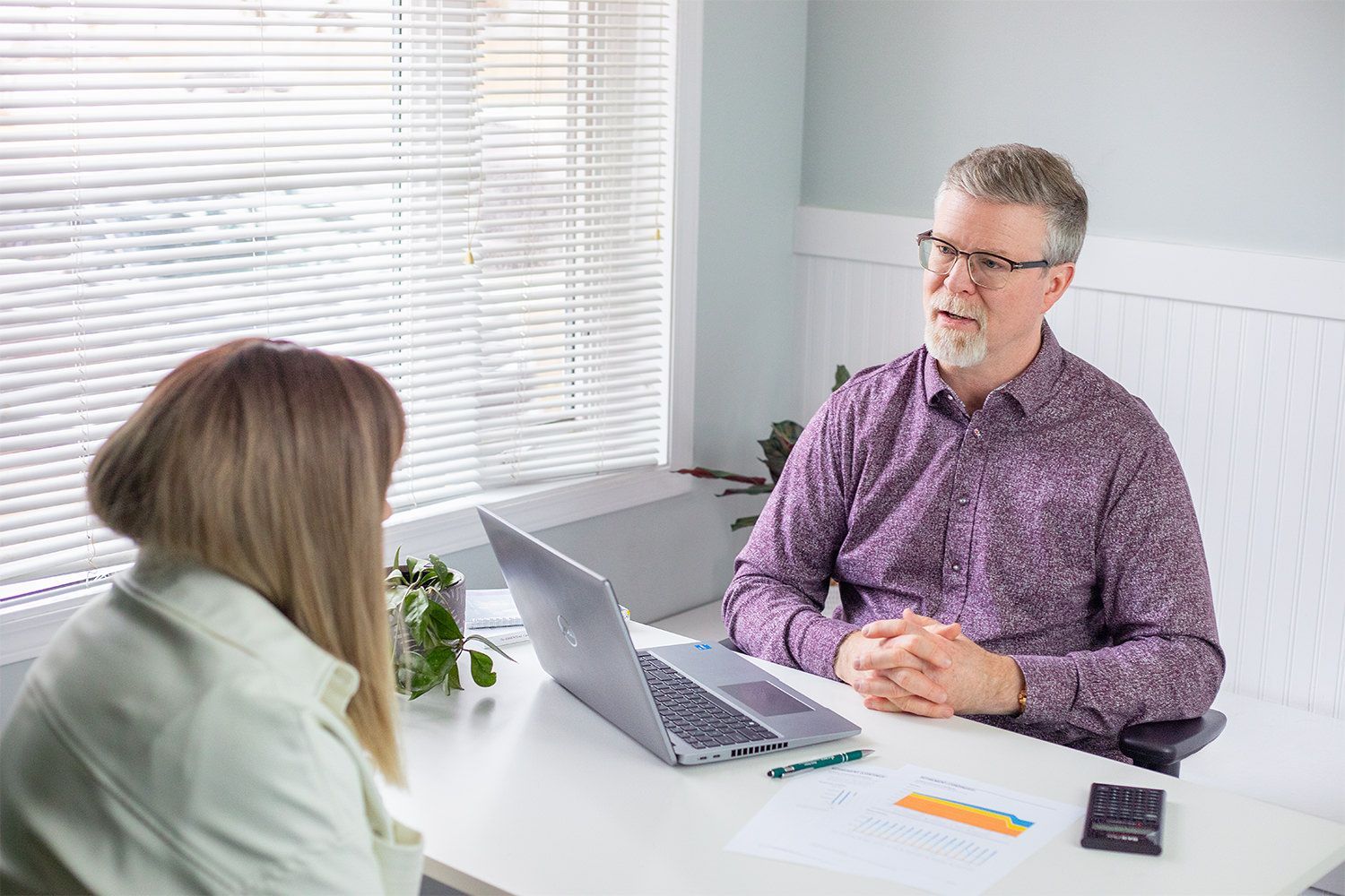 Woman and man at desk, laptop open. Man in purple shirt gestures, speaking. Window with blinds visible.