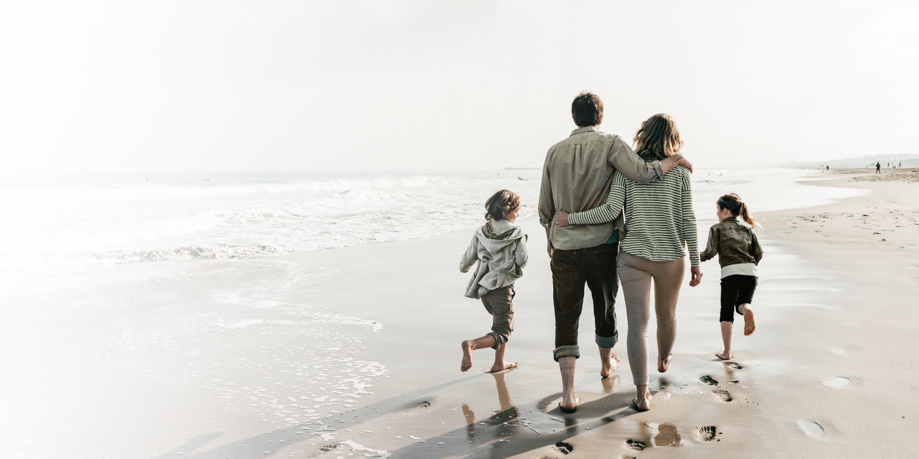 Family walking on a beach. A man and a woman have their arms around each other as they walk with two children toward the water.