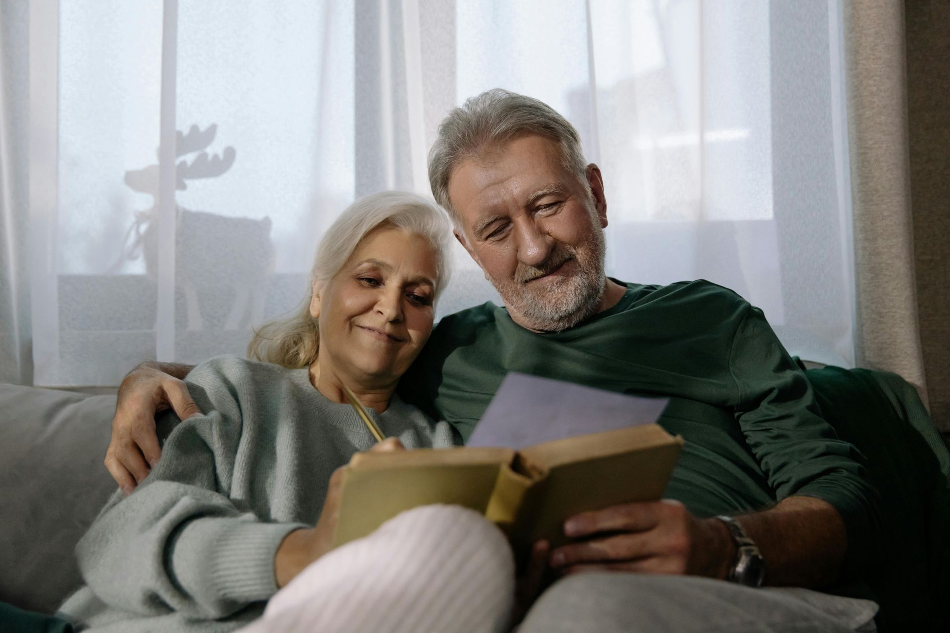 Elderly couple on a couch looking at a book, smiling. Man has arm around woman. Soft light, indoor setting.
