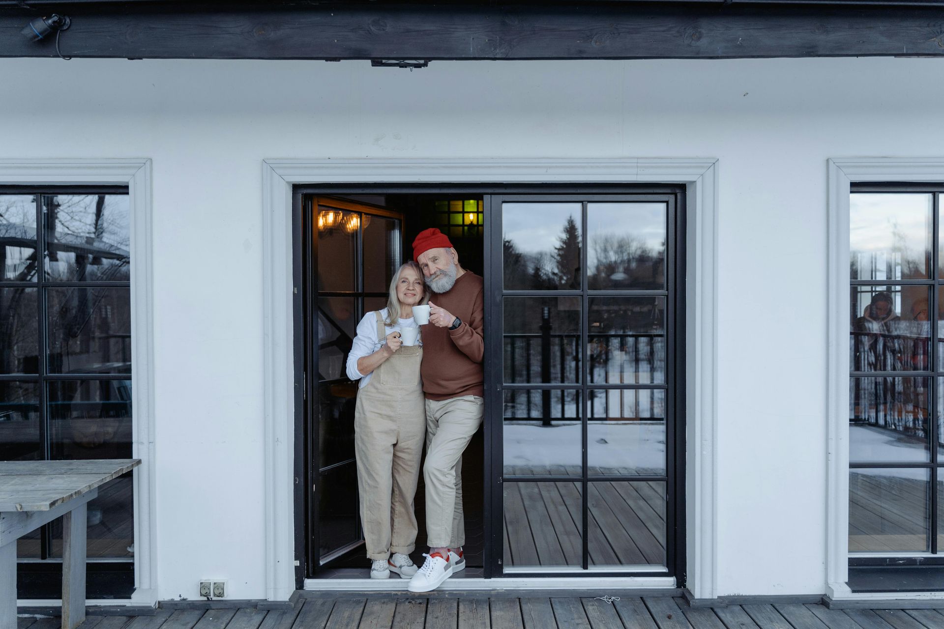 Couple standing in doorway, smiling, holding mugs. Snow covered surroundings.