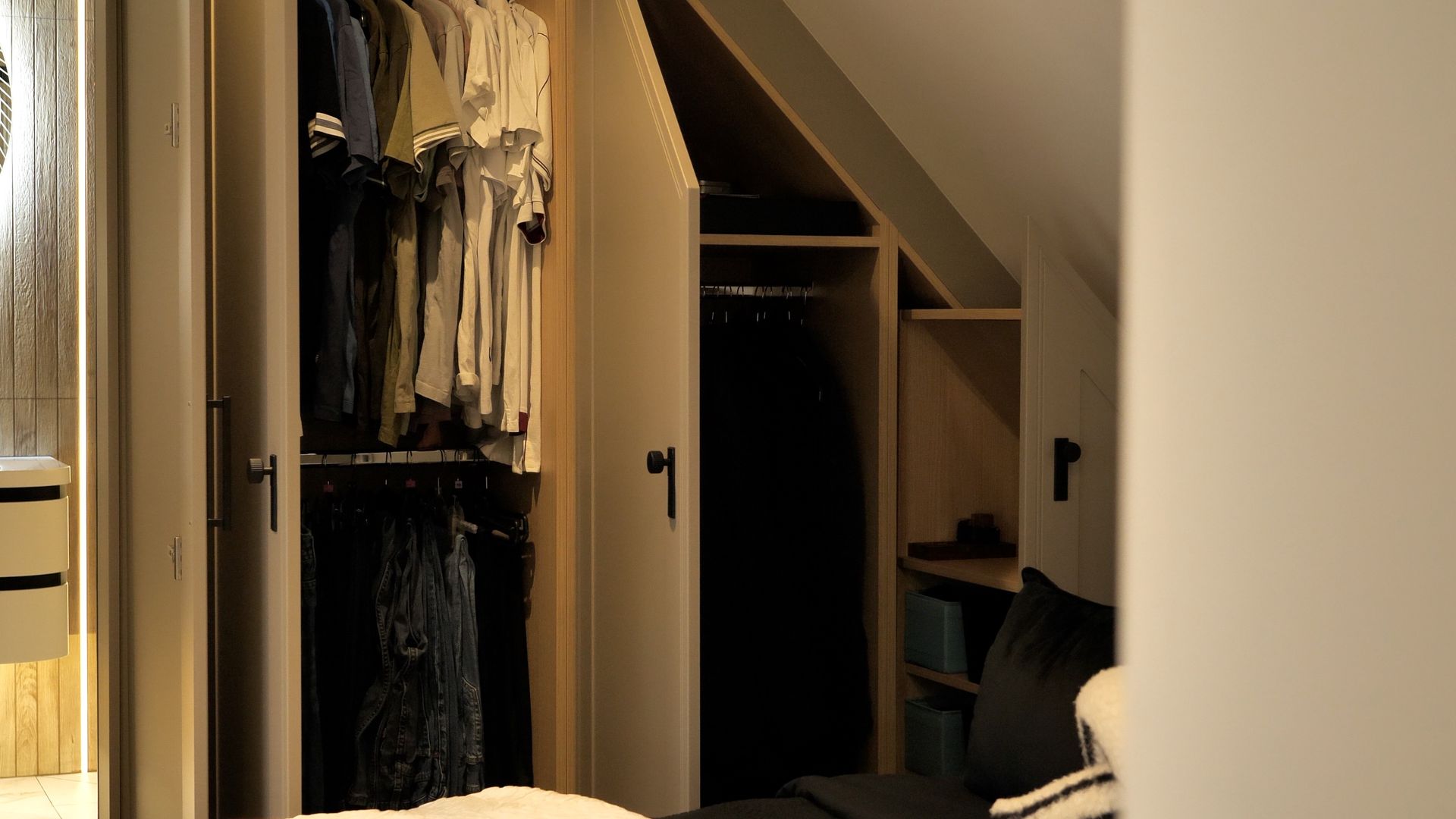 Closet with open doors, clothes hanging. Angled ceiling and shelves visible. Dark interior, neutral color scheme.