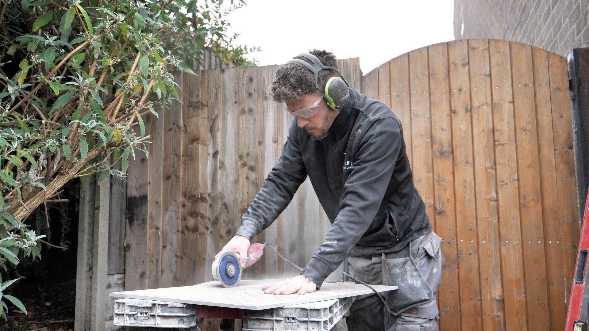 Man cutting tile with a power saw outside, wearing safety glasses and ear protection.
