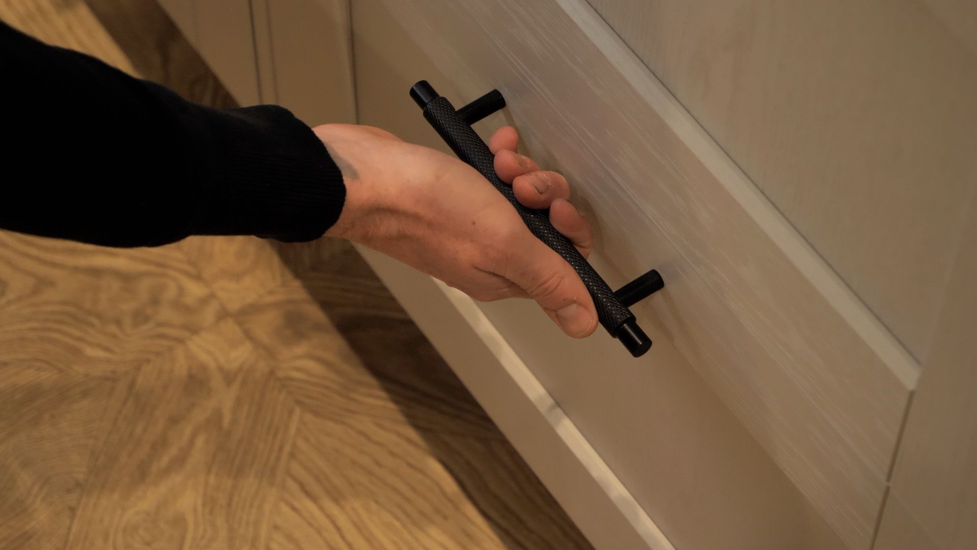 Hand pulling open a light-colored cabinet drawer with a black handle.
