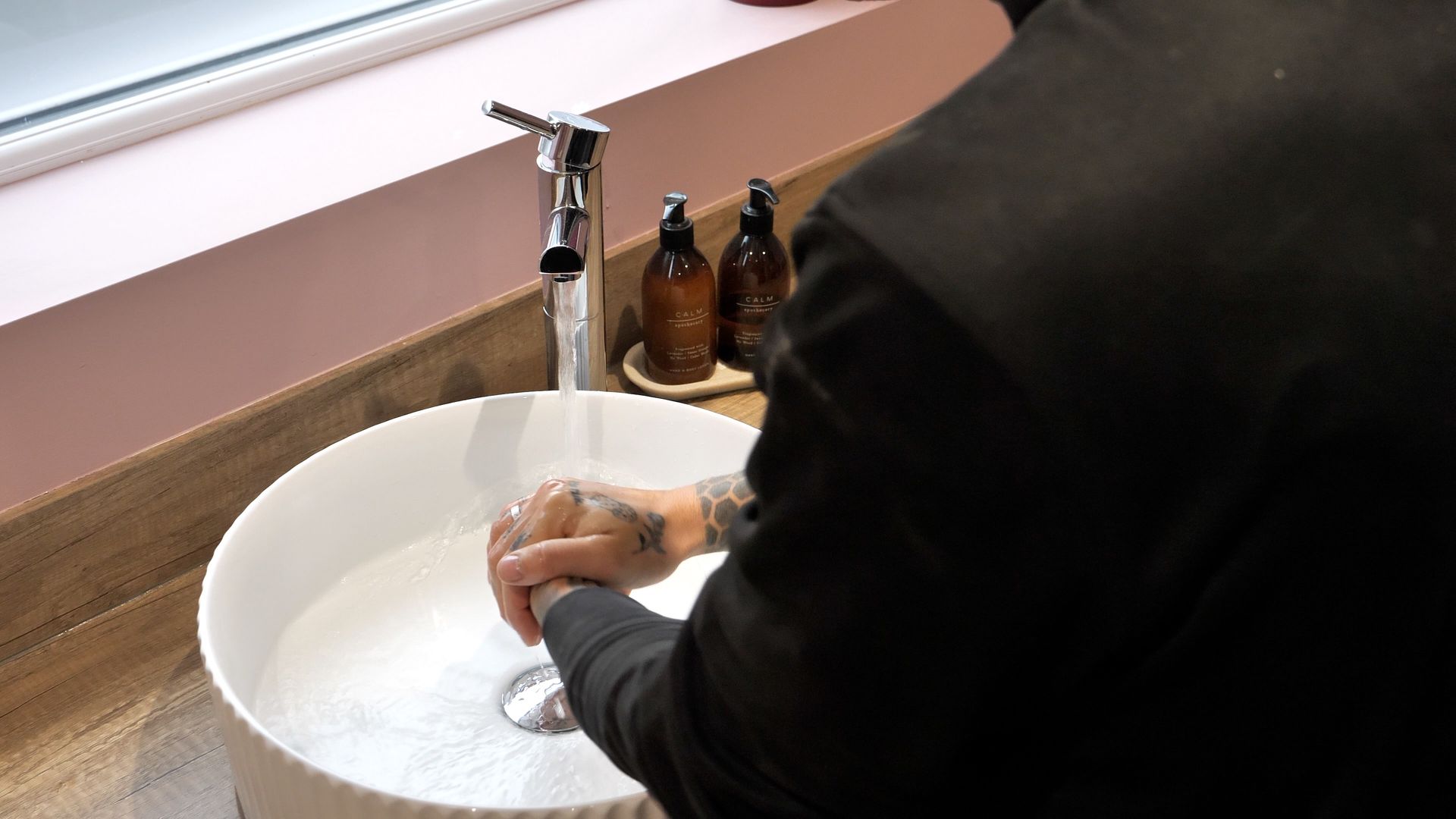 Person washing hands in a white sink under a chrome faucet. Light pink wall and brown countertop in the background.