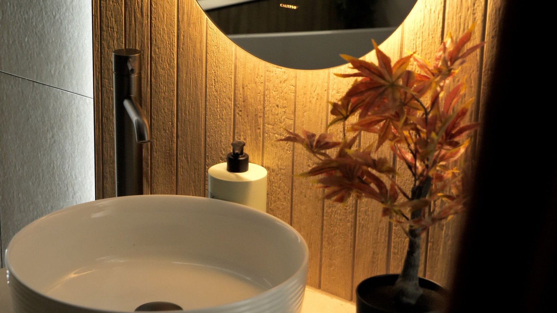 Bathroom sink with black faucet, round mirror, and decorative plant against a wood-paneled wall.