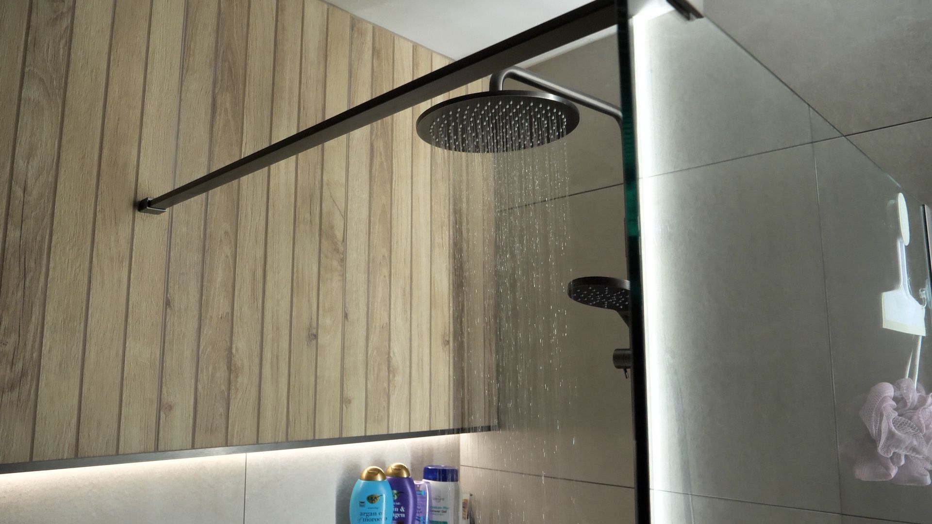 Shower with wood-paneled wall, glass door, and rain showerhead. Water streams down.