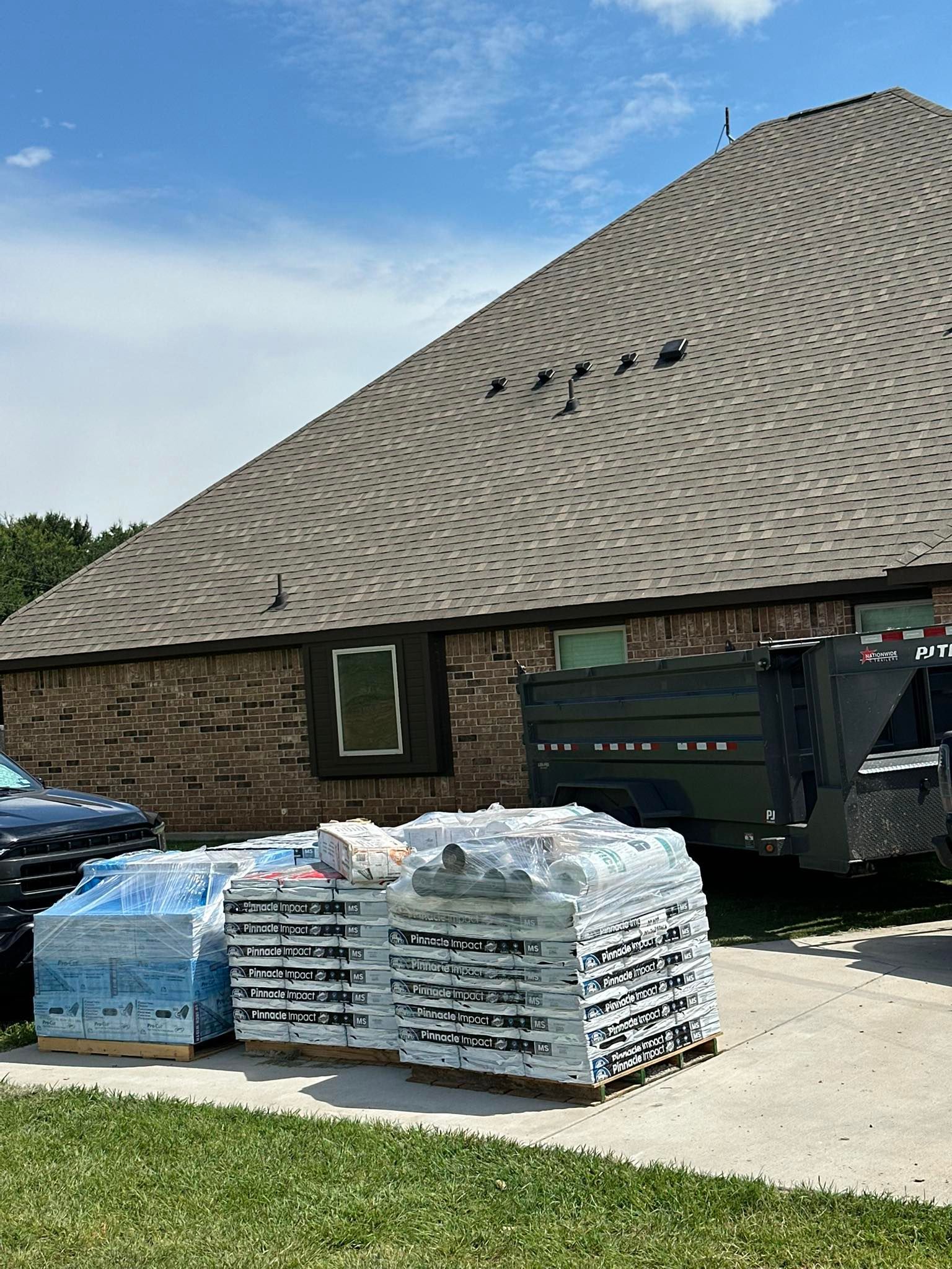 Pallets of bagged materials beside a brick building and green dumpster under a shingled roof.