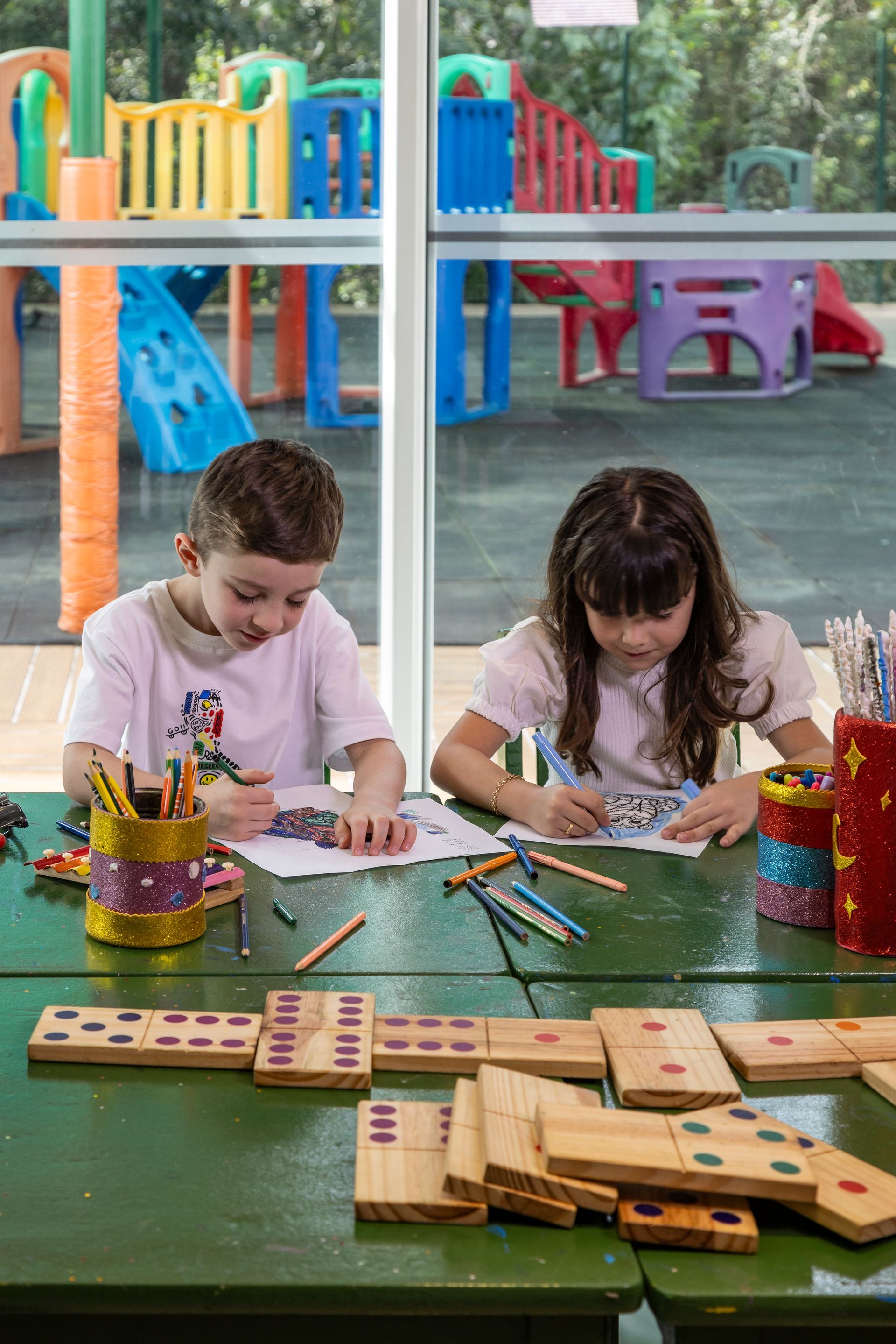 Um menino e uma menina estão sentados à mesa com dominós e lápis.