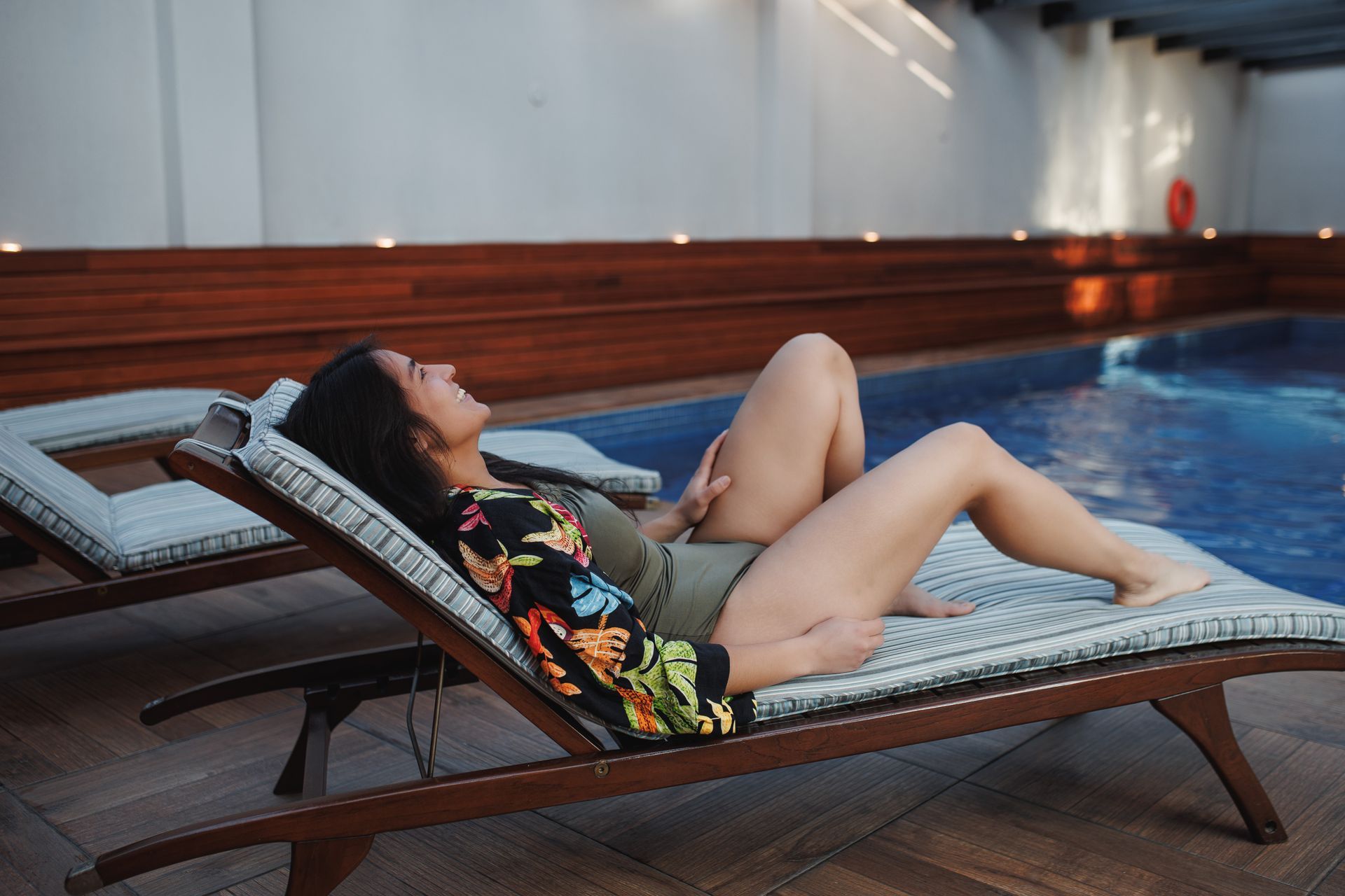 Mulher relaxando em uma espreguiçadeira ao lado de uma piscina. Ela está de maiô e rindo.