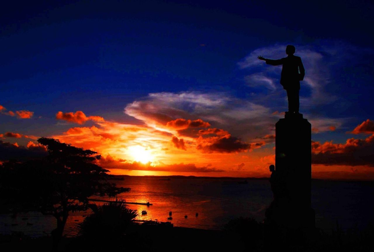 Silhouette of a statue pointing towards a vibrant sunset over the ocean; orange, blue, and yellow sky.