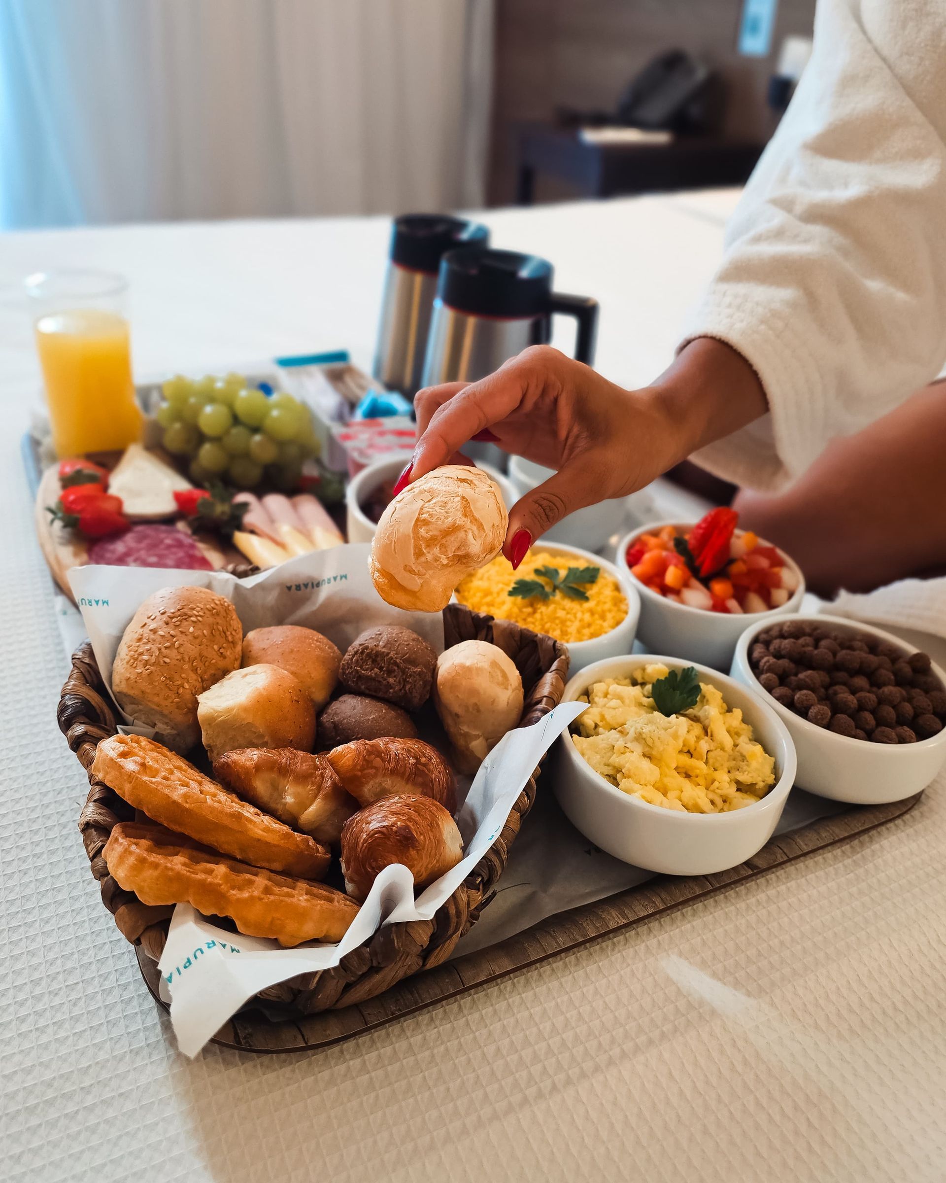 Breakfast tray on a bed: bread basket, scrambled eggs, fruit, juice, and coffee. A hand reaches for a roll.