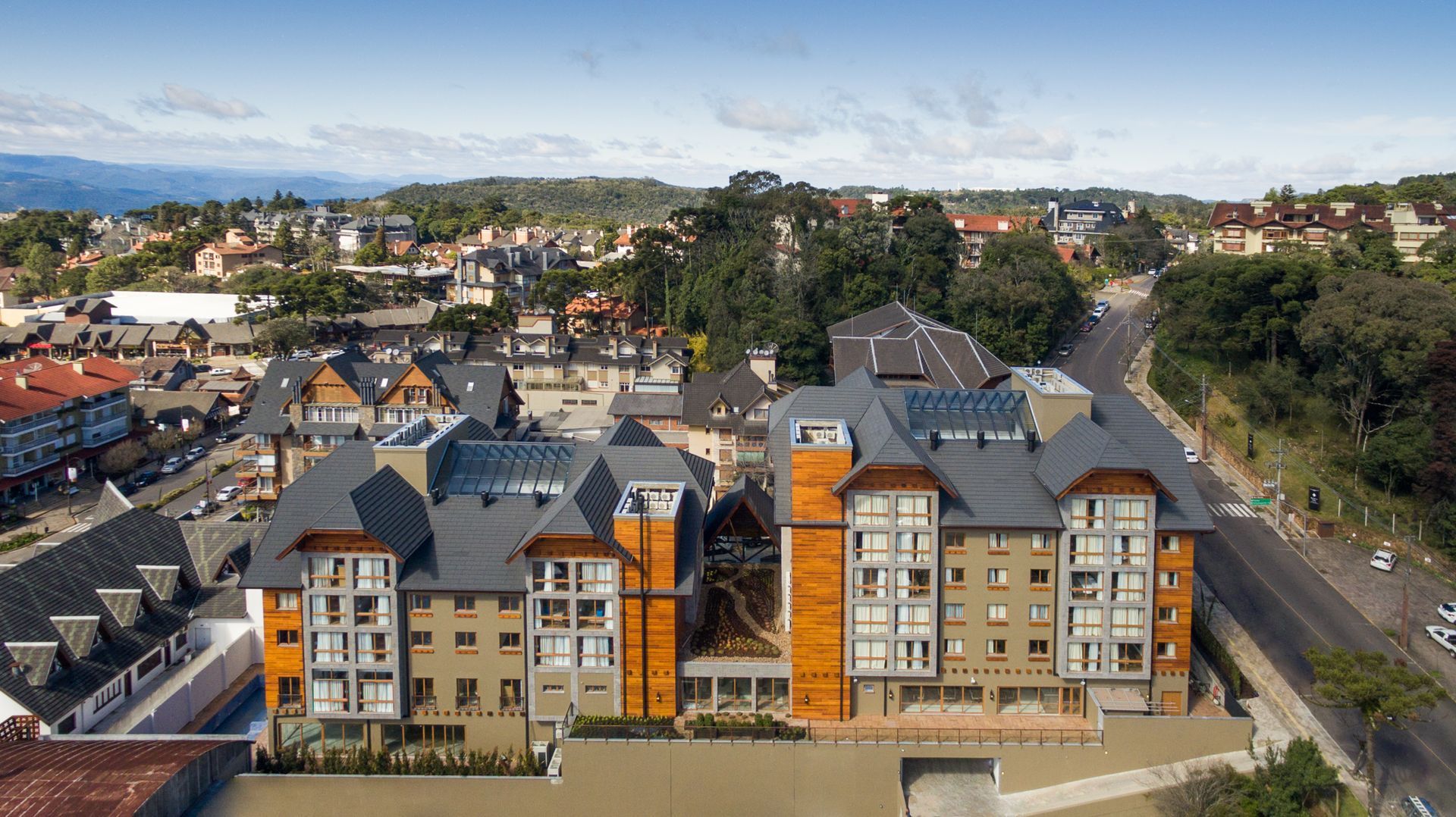 Aerial view of apartment buildings in a town, with gray roofs and tan siding. Road and green trees visible.