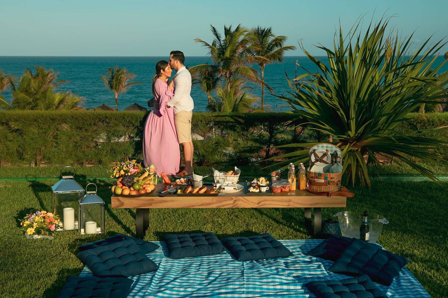 Casal se abraçando na beira do oceano, em um piquenique com comida e bebida, em um cobertor azul, em um dia ensolarado.