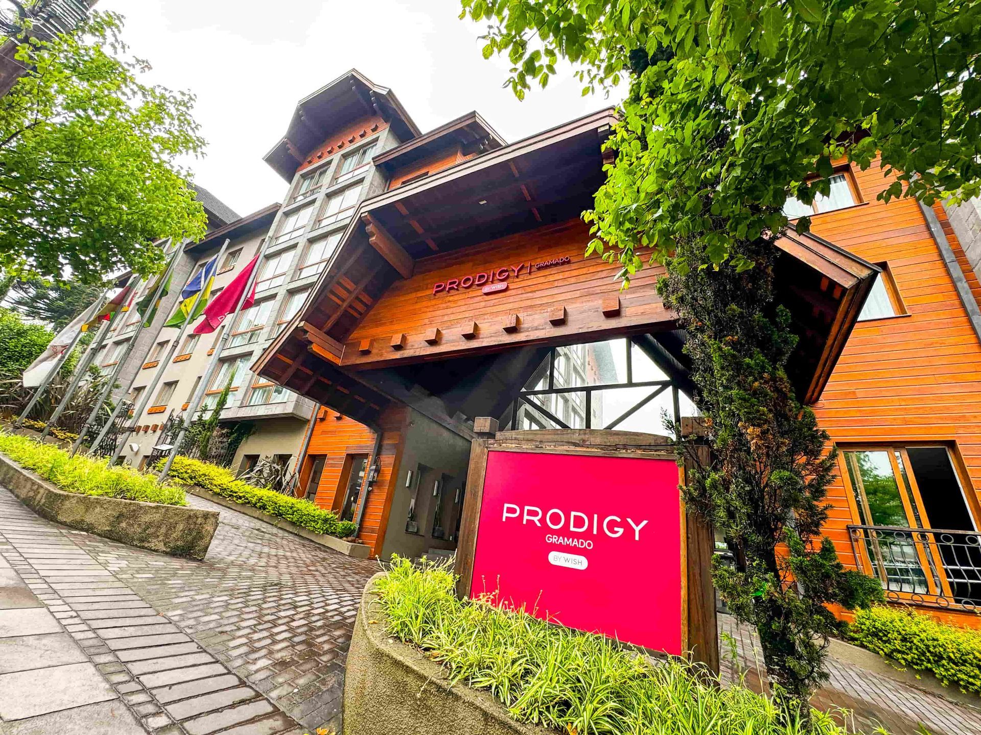 A bright pink sign for Prodigy Hotel stands in front of a multi-story, wood-paneled building on a stone pathway.