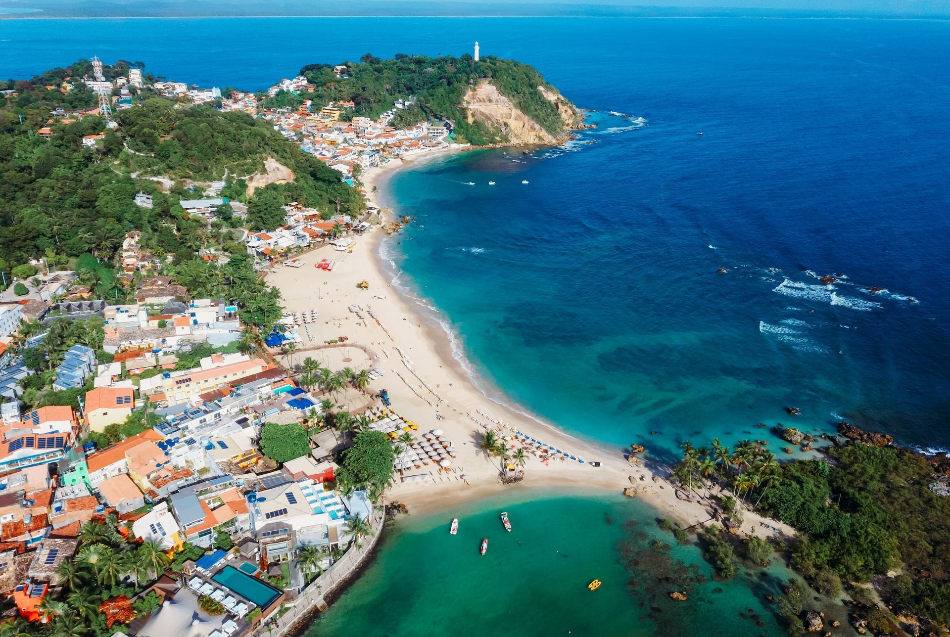 Aerial view of a tropical beach cove with turquoise water, white sand, and hillside houses.