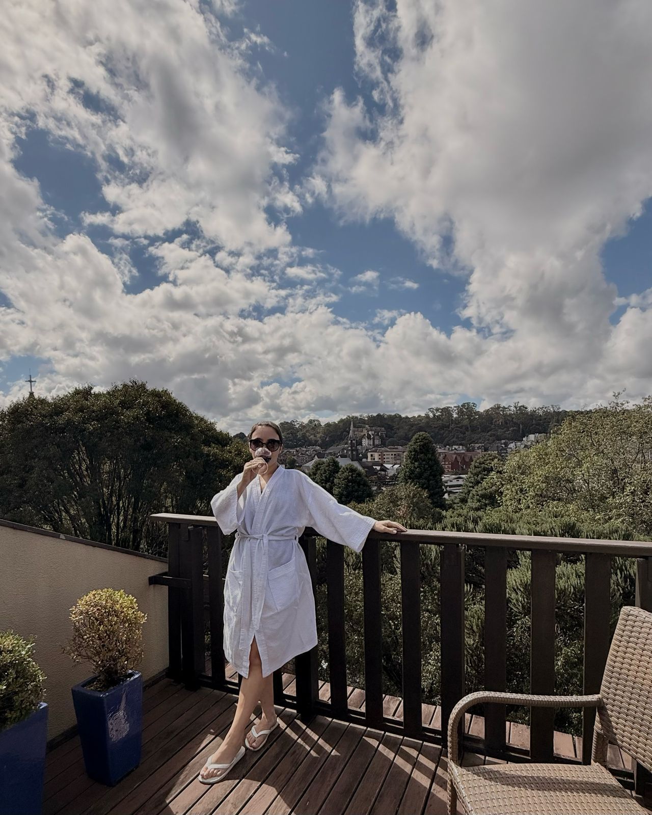 A woman in a bathrobe is standing on a balcony drinking wine.