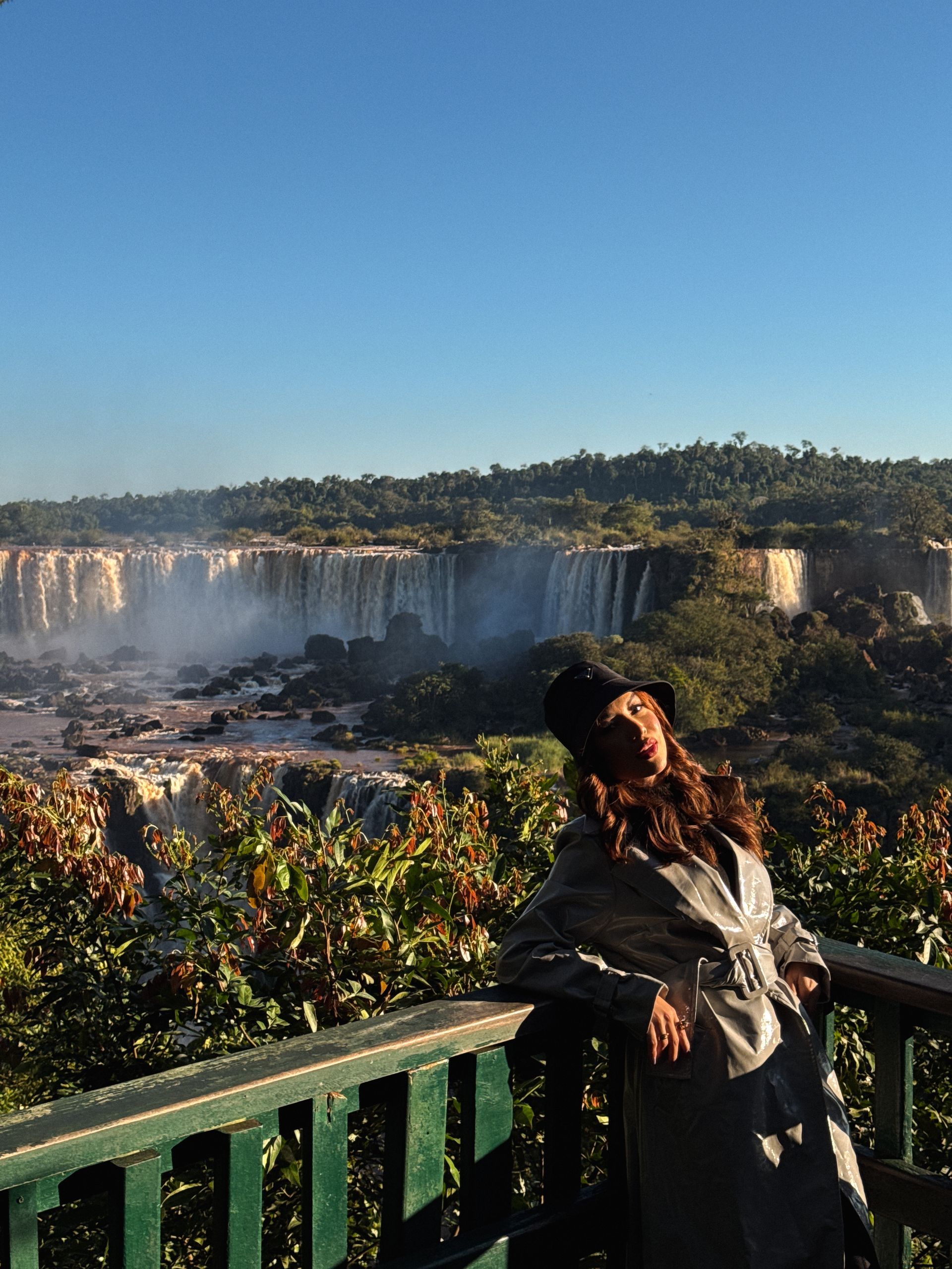 Woman in coat poses near Iguazu Falls, looking towards the camera. Lush greenery and blue sky.