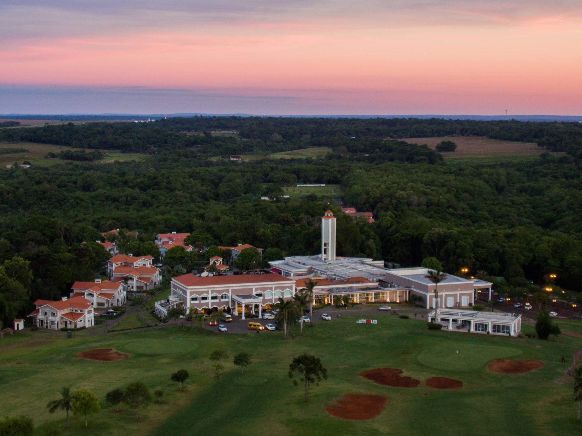 Aerial view of a resort with buildings, a golf course, and a forest at sunset.