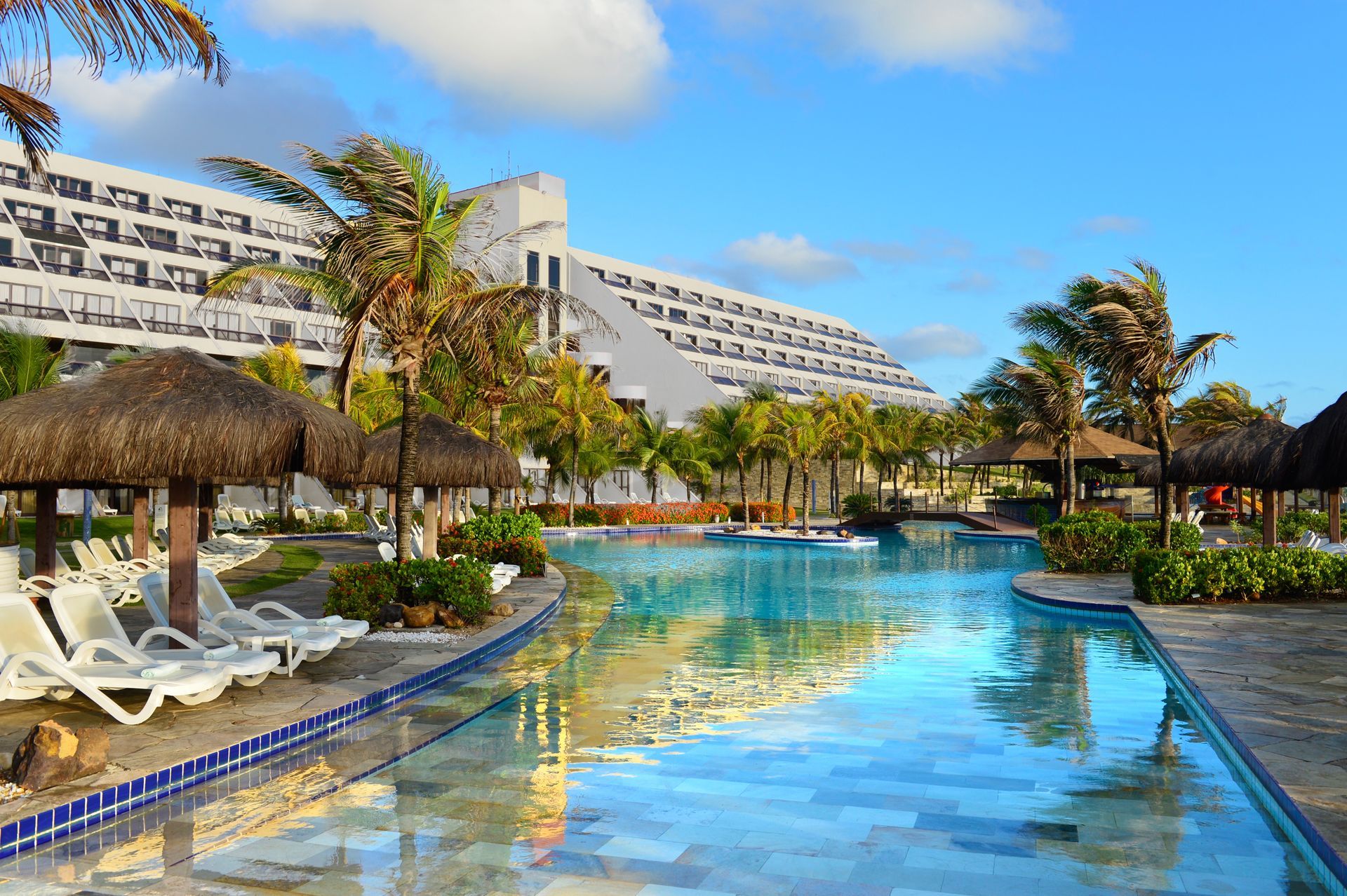 Pool with sun loungers, thatched umbrellas, palm trees, and a large resort building under a blue sky.