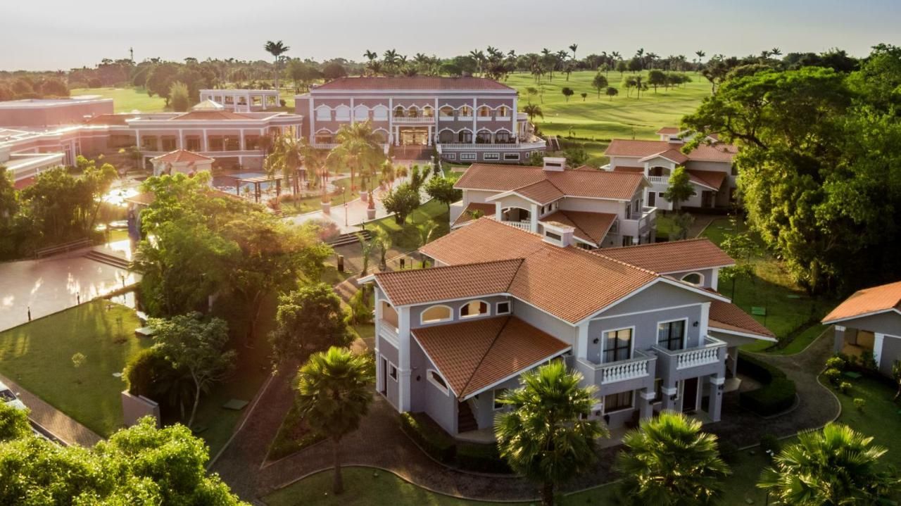Vista aérea de um resort bem iluminado, com edifícios de telhados vermelhos, gramados verdes e um campo de golfe, ao entardecer.