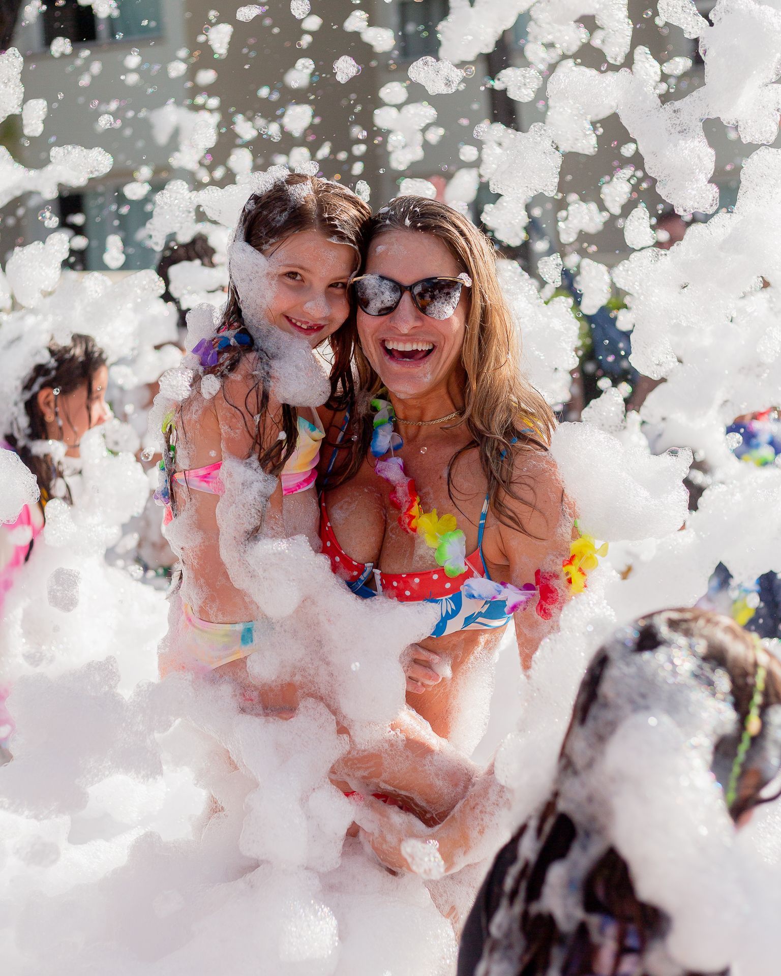 Woman holding child smiles in foam. Both are wearing swimwear and flower leis.