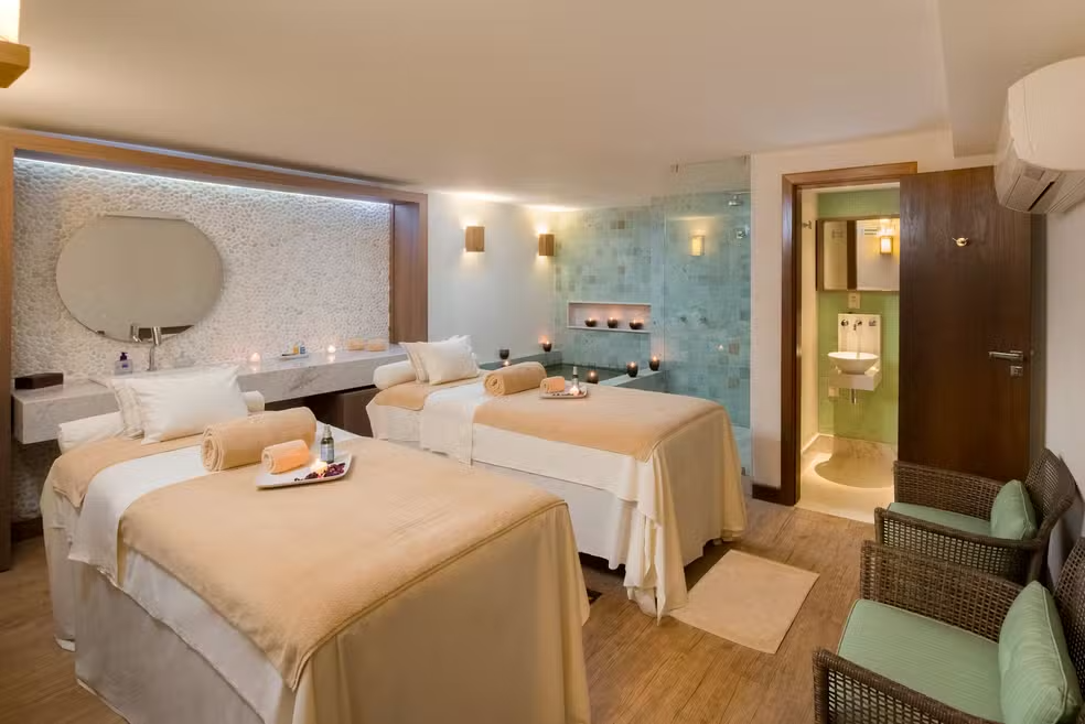 Spa room with two massage tables, sink, and chairs, soft lighting, and a bathroom.