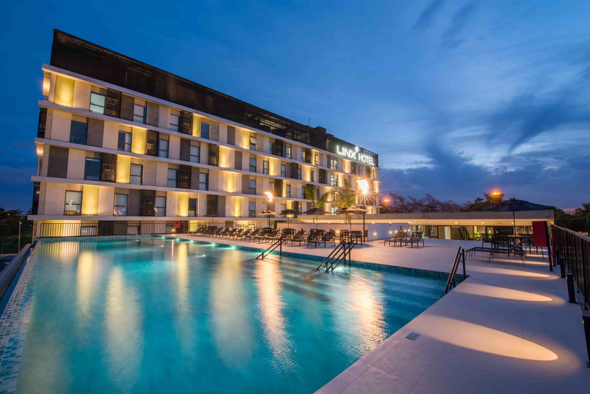 Hotel moderno com varandas iluminadas junto a uma piscina turquesa ao entardecer.