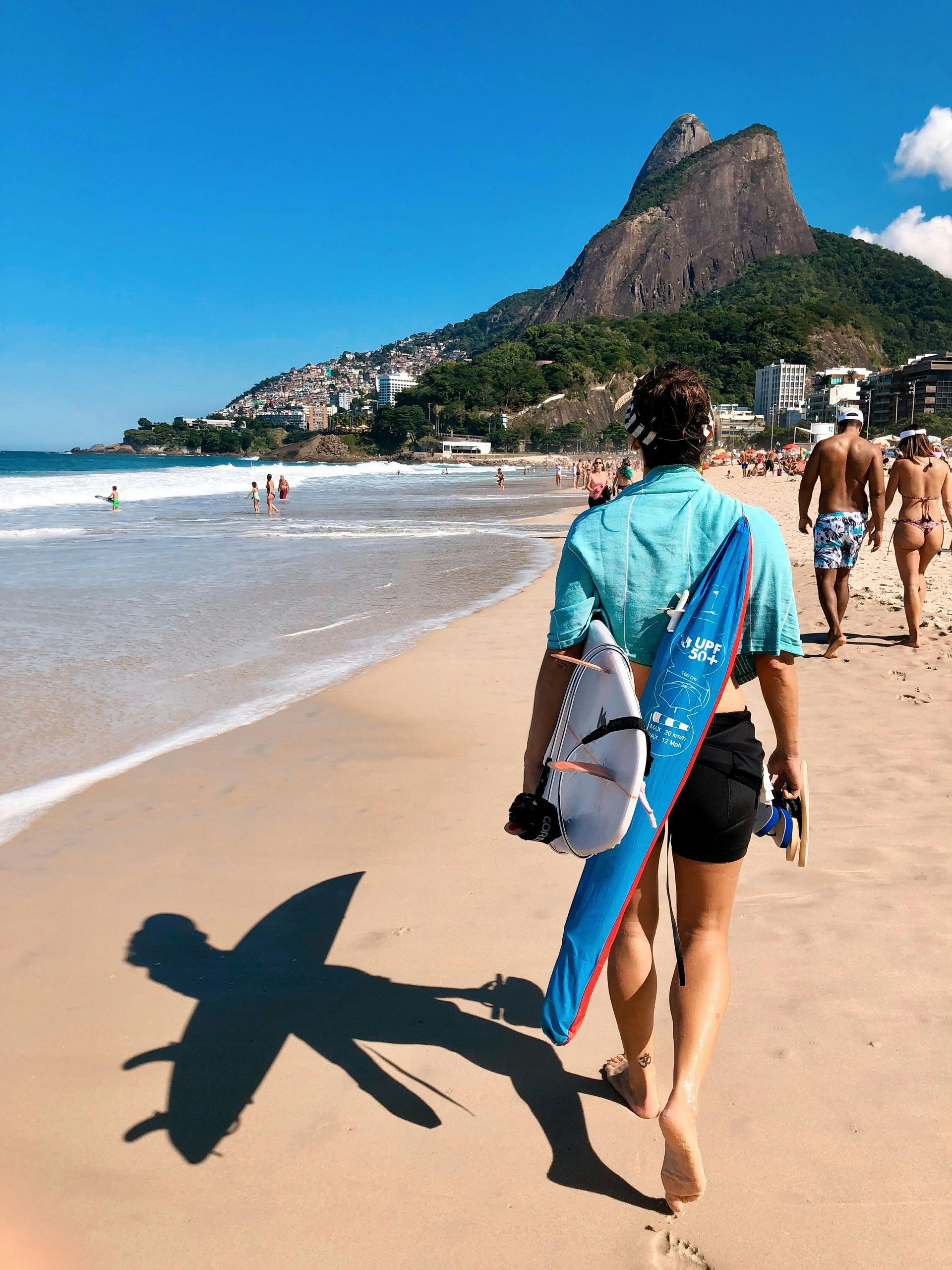 Homem caminha na praia com uma prancha de surfe, Praia de Ipanema, Rio de Janeiro, com o Pão de Açúcar ao fundo. Dia ensolarado.