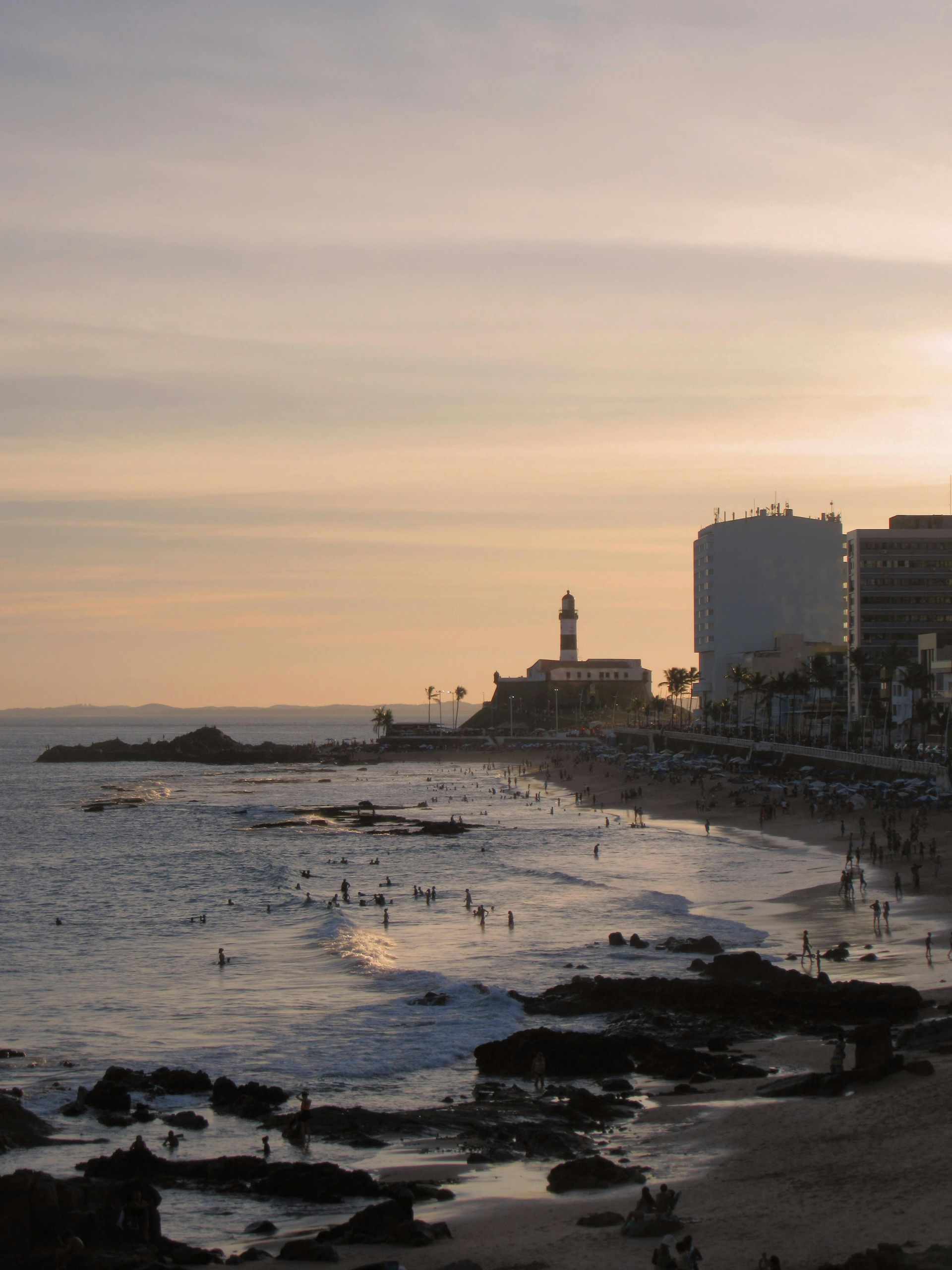 Cena costeira ao pôr do sol, ondas do oceano, praia com pessoas, edifícios e um farol sob um céu dourado.