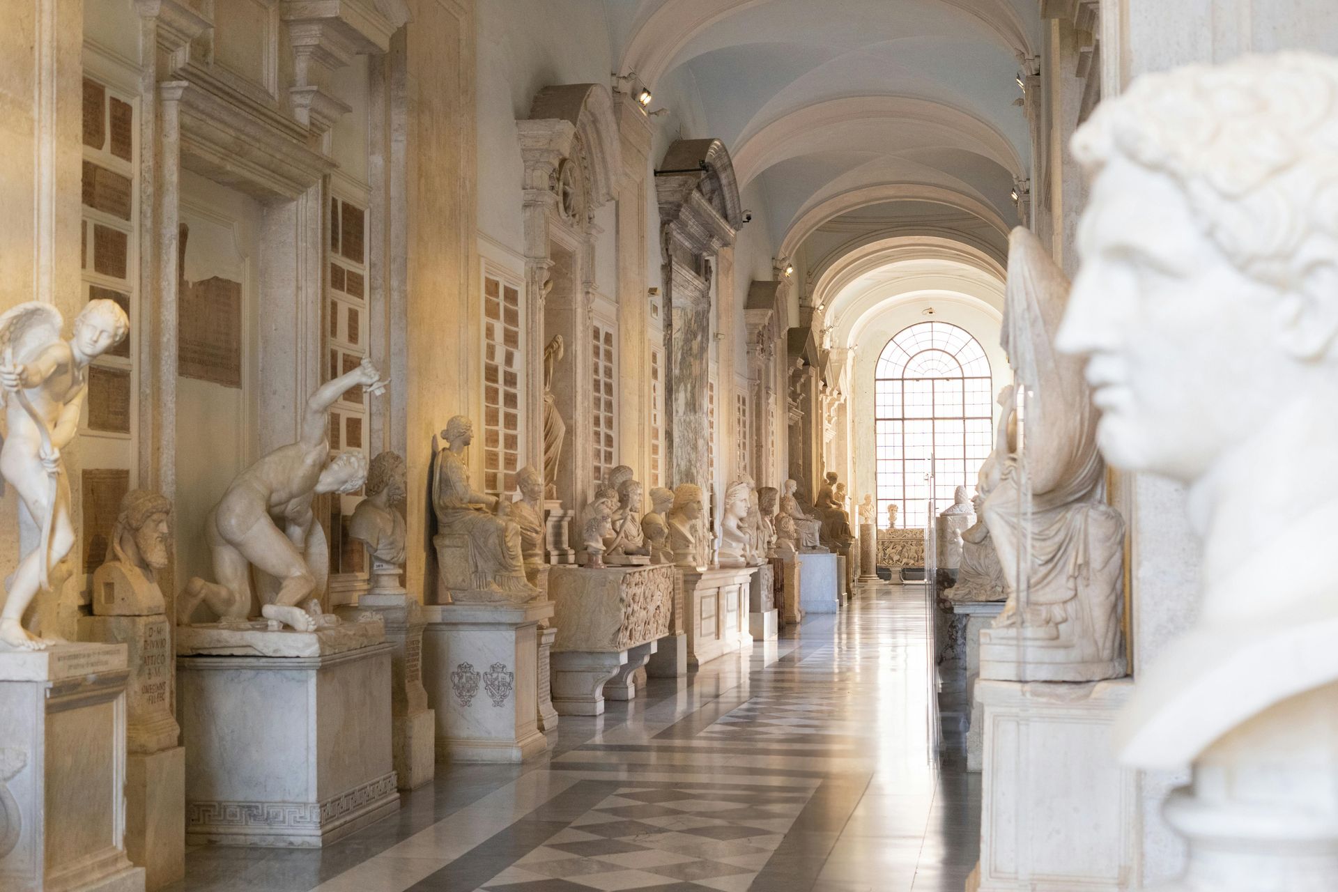 Hallway in a museum lined with marble sculptures and busts, natural light streaming from a large window.