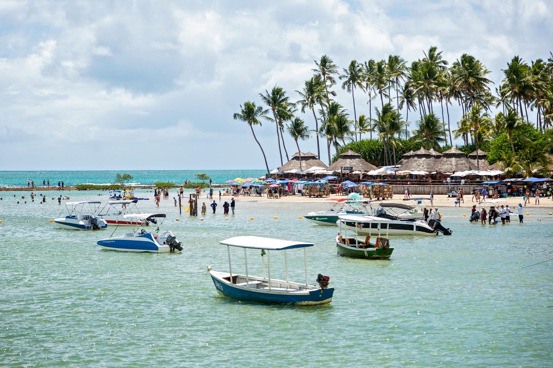Sunny tropical beach with anchored boats, swimmers, and palm trees along a turquoise shore