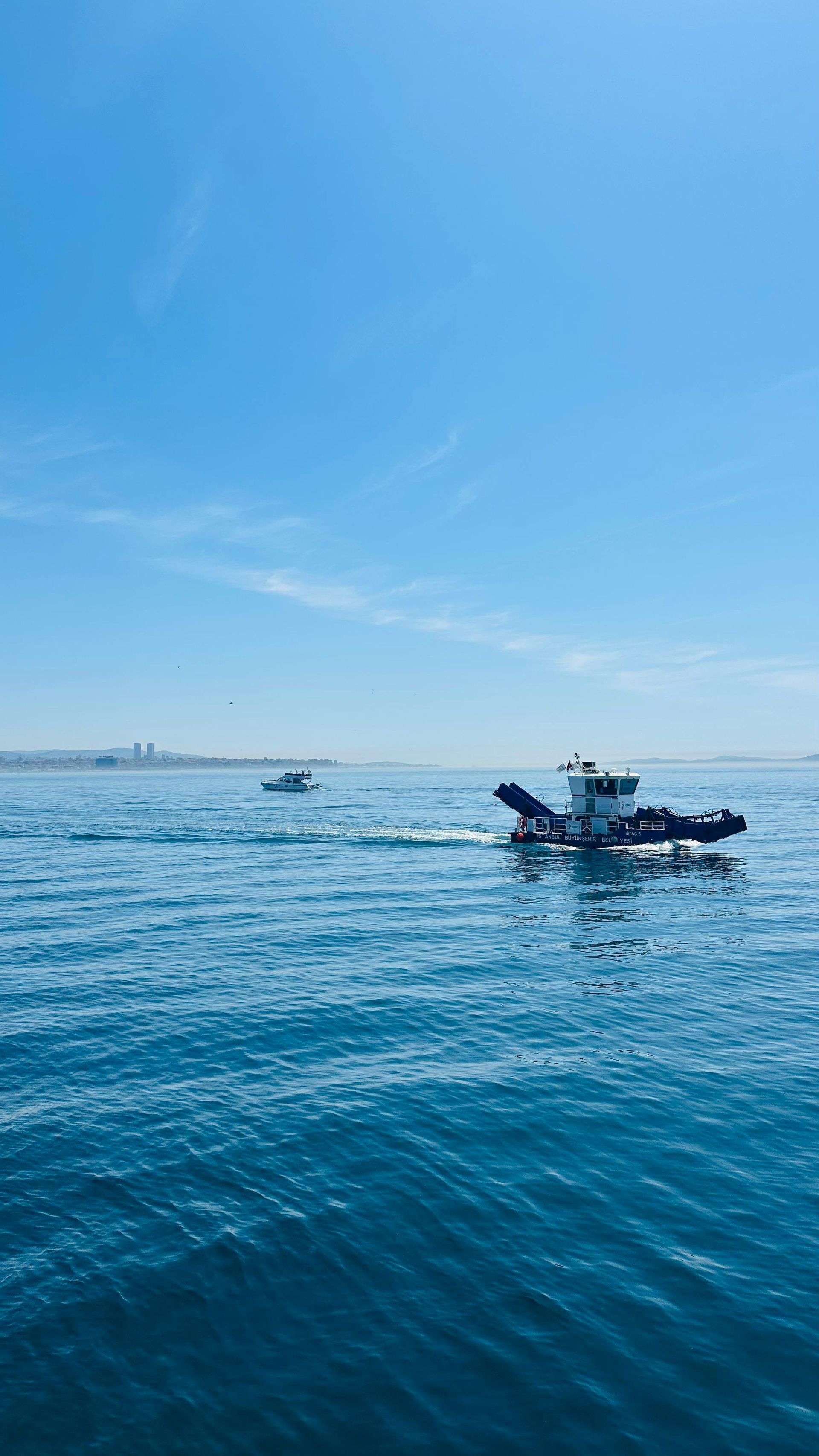 Um pequeno barco de trabalho navega pelas águas calmas e azul-claras do oceano sob um céu limpo, com um navio distante no horizonte.