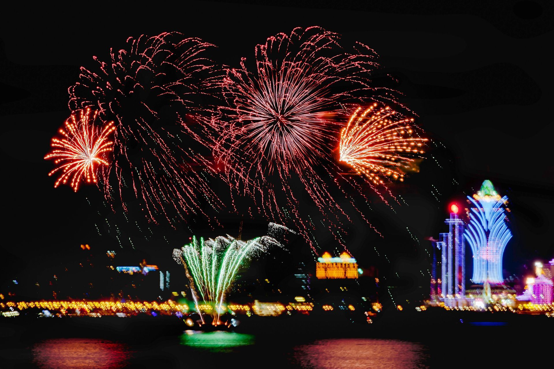 Fireworks over a waterfront at night, featuring red, green, and gold bursts and illuminated buildings.